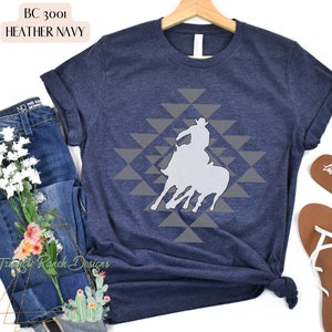 Pu&ograve; includere: Una t-shirt blu navy heather con una silhouette bianca di un cowboy a cavallo su uno schema geometrico grigio. La t-shirt &egrave; realizzata in un materiale morbido e confortevole.