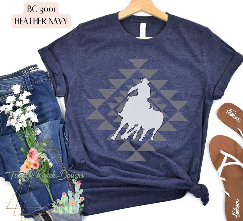 Pu&ograve; includere: Una t-shirt blu navy heather con una silhouette bianca di un cowboy a cavallo su uno schema geometrico grigio. La t-shirt &egrave; realizzata in un materiale morbido e confortevole.