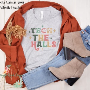 May include: A gray t-shirt with the text "TECH THE HALLS" in colorful retro-style lettering. The shirt is styled with a brown jacket, blue jeans, and brown ankle boots.