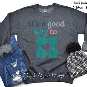 K&ouml;nnte beinhalten: Dunkelgraues Sweatshirt mit dem Text "it's a good day to CODE" in T&uuml;rkis, Rosa und Grau. Das Sweatshirt wird mit einer blauen Jeans und einer schwarz-wei&szlig; gestrickten M&uuml;tze mit einem schwarzen Bommel pr&auml;sentiert. Der Markenname "Triangle Ranch Designs" steht unten.