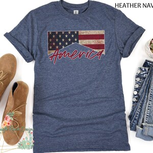 May include: A heather navy blue t-shirt with a distressed American flag design and the word "America" written in red cursive.