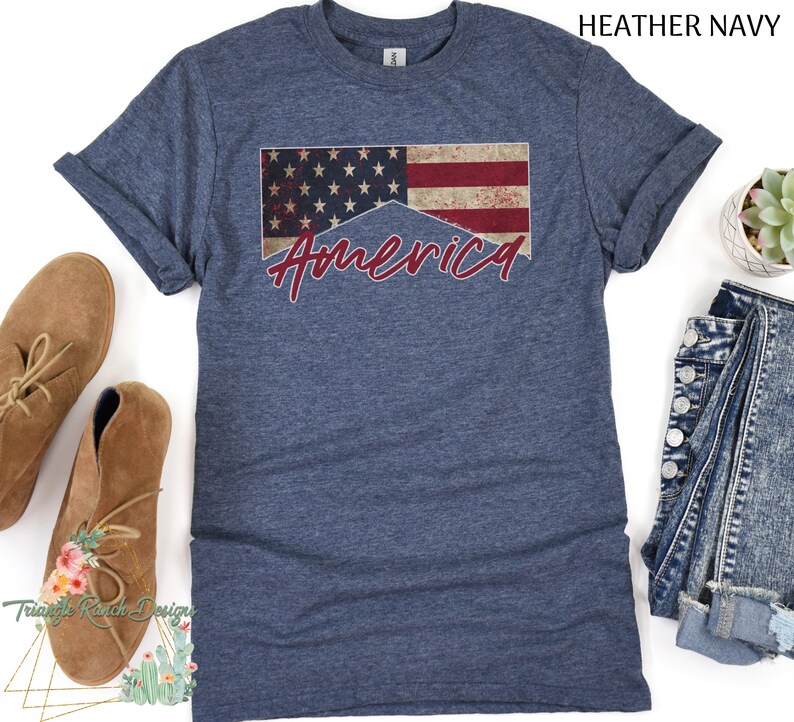 May include: A heather navy blue t-shirt with a distressed American flag design and the word "America" written in red cursive.