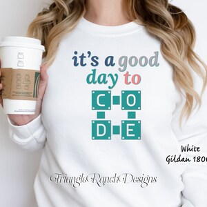 K&ouml;nnte beinhalten: Wei&szlig;es Gildan 18000 Sweatshirt mit dem Text "it's a good day to CODE" in T&uuml;rkis, Rosa und Blau. Das Design zeigt eine quadratische Grafik mit den Buchstaben C, O, D und E. Eine Kaffeetasse wird im Bild gehalten.