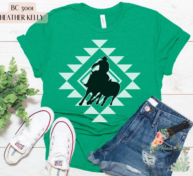Pu&ograve; includere: Una maglietta verde con un design geometrico bianco che presenta la silhouette di un cowboy che cavalca un toro. Il testo "BC 3001 HEATHER KELLY" &egrave; in cima alla maglietta.