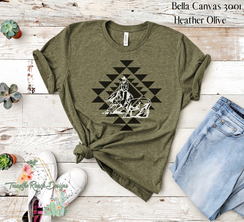 May include: A green Bella Canvas 3001 t-shirt with a white graphic of a cowgirl riding a horse and a steer in a geometric pattern. The text "Heather Olive" is visible in the top right corner of the image.