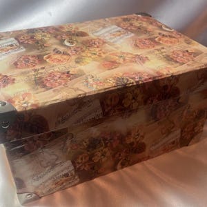 Puede incluir: Una caja decorativa de estilo vintage con un diseño floral y notas musicales. La caja tiene esquinas metálicas y una tapa con bisagras.