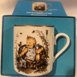 Può includere: Una tazza da bambino in porcellana bianca con un bordo dorato. La tazza presenta un'illustrazione colorata di un bambino seduto in un cesto con un uccello e dei fiori. Il testo "1973 Child's Cup Porcelain" è stampato sulla scatola.