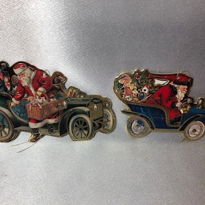Könnte beinhalten: Zwei Vintage-Weihnachtsschmuckstücke in Form von Oldtimern. Eines ist dunkelgrün, das andere blau. Beide zeigen den Weihnachtsmann am Steuer, mit Passagieren und Geschenken. Goldener Rand umrandet die Ornamente.
