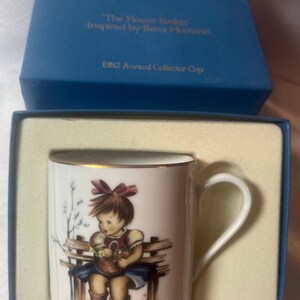 Può includere: Tazza in ceramica bianca con bordo dorato, raffigurante una ragazza con un cestino di fiori seduta su una panchina. La tazza è all'interno di una scatola blu con il testo "The Flower Basket" e "1982 Annual Collector Cup".
