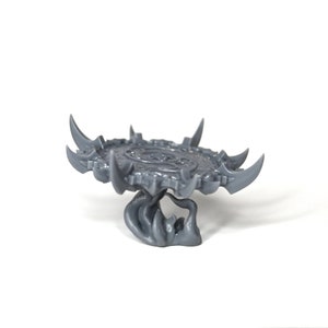 May include: A gray plastic miniature of a circular platform with a detailed design and sharp points around the edge. The platform is supported by a gray plastic base with a textured, organic design.