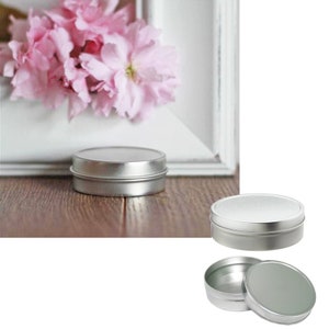 May include: Two silver metal tins with lids. The tin on the left is closed. The tin on the right is open, showing the inside of the tin. The lid is sitting next to the tin.