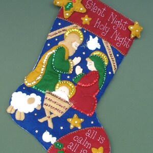 May include: A blue felt Christmas stocking with a red felt top and a scene of the nativity. The stocking has gold glitter stars and sequins, and the text "Silent Night Holy Night all is calm all is bright".