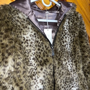 May include: A brown and white leopard print faux fur jacket with a black zipper and a satin lining. The jacket has a hood and is hanging on a white hanger.