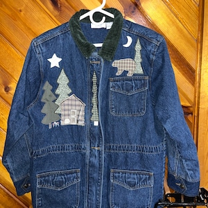 May include: A blue denim jacket with a corduroy collar and appliques of a cabin, trees, a bear, a star, and a crescent moon.