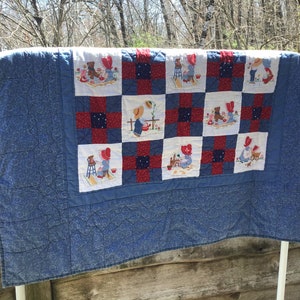 May include: A blue and red patchwork quilt with embroidered squares featuring children and teddy bears at play. The quilt has a blue patterned border and is displayed outdoors, draped over a white rod.
