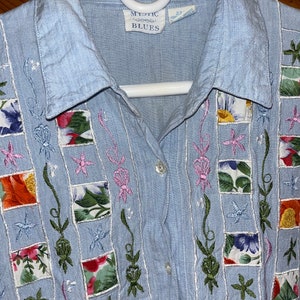 May include: Light blue button-down shirt with floral patchwork squares and embroidered accents. The shirt features the text "Mystic Blues" on the label. The shirt is size 18-22.