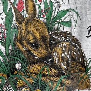 May include: A close-up of a gray t-shirt featuring a detailed illustration of a fawn nestled among green grass, red and white flowers, and brown leaves. The fawn has brown fur with white spots and large, dark eyes. The design has a vintage, nature-inspired aesthetic.