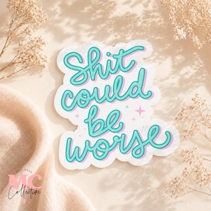 May include: A white sticker with the phrase "Shit could be worse" in teal script with a light pink outline. The sticker has a white border and is surrounded by dried flowers.