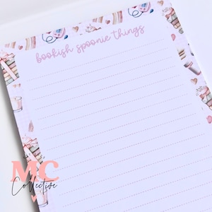 May include: A notepad with the words "bookish spoonie things" in pink script. The notepad has a pale pink background with a border featuring books, teacups, and hearts. The bottom left corner has the text "MC Collective".
