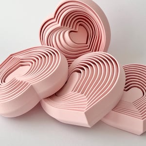 May include: A collection of four light pink heart-shaped objects, each featuring a series of concentric, stepped layers that create a three-dimensional, ridged effect. The objects are arranged in a cluster, showcasing their smooth, matte finish.