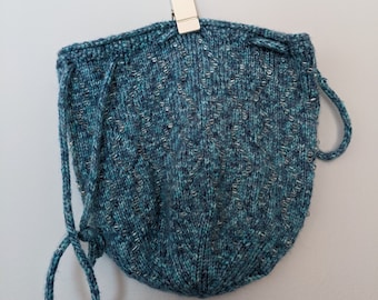 May include: A teal blue knitted drawstring bag with a beaded pattern. The bag is hanging from a white wall with a silver clip.