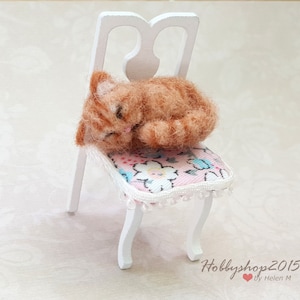 May include: A small, orange and white cat figurine is curled up and sleeping on a white chair with a pink and white floral cushion. The chair has a white, ornate back and legs.