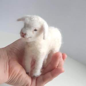 Puede incluir: Figura de cordero de lana blanca hecha a mano. El cordero tiene un aspecto esponjoso y texturizado con una nariz rosada y pequeños detalles. El cordero se sostiene en una mano, mostrando su tamaño y artesanía. Una decoración encantadora y única.