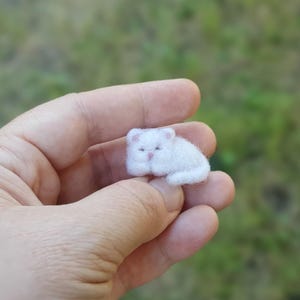 Puede incluir: Una figura miniatura de gato hecha de lana fieltrada blanca, con una textura suave y esponjosa, orejas pequeñas y puntiagudas y una delicada nariz rosa, representada en una posición de descanso acurrucada con la cola pegada al cuerpo.