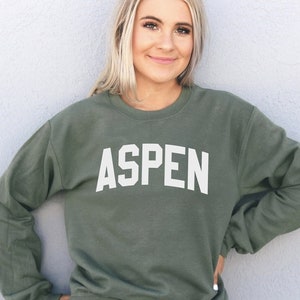 May include: A woman wearing a green sweatshirt with the word "ASPEN" printed in white on the front. She is standing in front of a white wall.