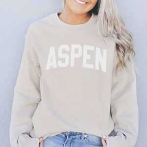 May include: A beige sweatshirt with the word "ASPEN" printed in white on the front. The model is wearing blue jeans.