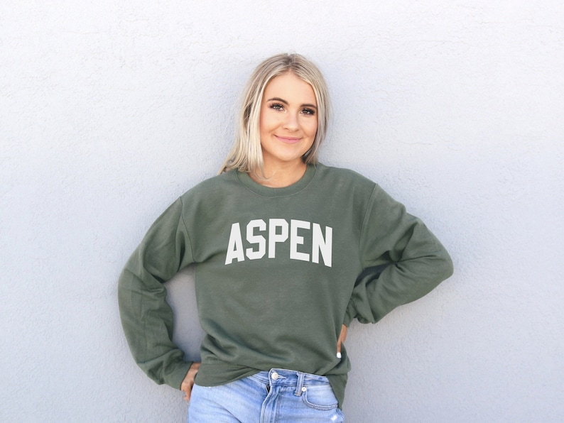 May include: A woman wearing a green sweatshirt with the word "ASPEN" printed in white on the front. She is standing in front of a white wall.