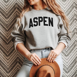 May include: A gray sweatshirt with the word "ASPEN" printed in black block letters. The sweatshirt is being worn by a person with long blonde hair. The person is holding a brown hat in their hands.