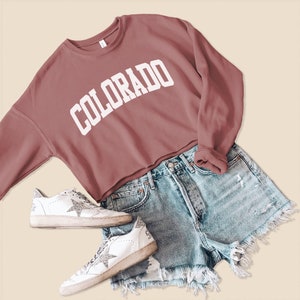 May include: A cropped sweatshirt in a dusty rose color with the word "COLORADO" printed in white on the front. The sweatshirt is paired with denim shorts and white sneakers.