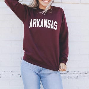 A woman wearing a burgundy sweatshirt with the word "ARKANSAS" printed on it, standing in front of a white brick wall.