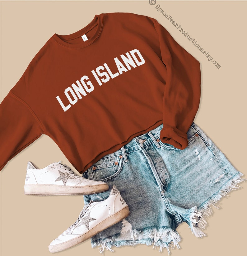 Pu&ograve; includere: Una felpa corta color ruggine con la scritta "LONG ISLAND" in lettere bianche. La felpa &egrave; abbinata a pantaloncini di jeans blu chiaro e scarpe da ginnastica bianche.