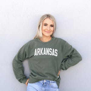 A smiling woman wearing a green sweatshirt with the word "ARKANSAS" printed on it, standing against a white wall.