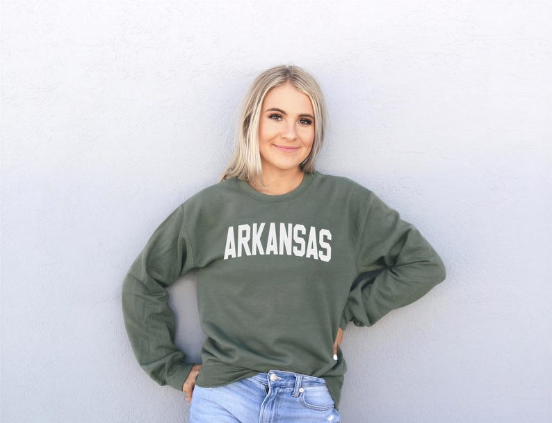A smiling woman wearing a green sweatshirt with the word "ARKANSAS" printed on it, standing against a white wall.