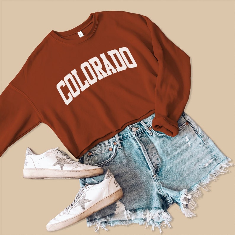 May include: A rust-colored cropped sweatshirt with the word "COLORADO" in white lettering. The sweatshirt is paired with light blue denim shorts and white sneakers with silver glitter.