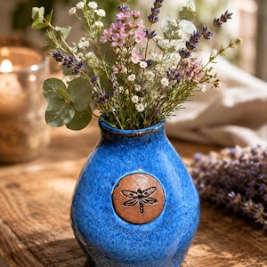 May include: A vibrant blue ceramic vase with a glossy, textured finish and a round, dark brown clay medallion featuring an embossed dragonfly design. The vase is filled with a bouquet of lavender, eucalyptus, and small white flowers.