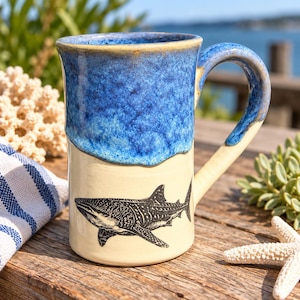 May include: A ceramic mug featuring a two-tone design with a glossy blue glaze on the upper portion and a cream-colored base. A detailed black ink illustration of a whale shark is printed on the lower cream section of the mug.