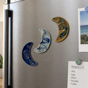 May include: Three crescent moon-shaped ceramic magnets with spiral patterns. One magnet is dark blue, one is light blue with white streaks, and one is a golden-brown color. They are attached to a metallic surface near a shopping list note.