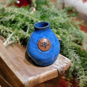 May include: A small, blue ceramic vase with a dragonfly design. The vase is sitting on a wooden surface.