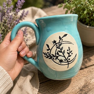 Puede incluir: Una taza de cerámica turquesa con una inserción circular grande de color crema en la parte frontal. La inserción presenta un diseño negro grabado de una luna creciente, estrellas pequeñas y una rama con hojas. Tiene un asa curva.