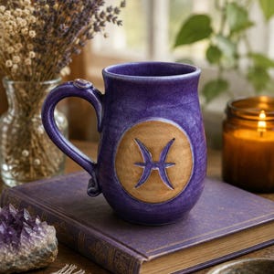 May include: A purple ceramic mug featuring a circular tan medallion on the front with the Pisces zodiac symbol embossed in purple. The mug has a curved handle and a smooth, glossy finish, showcasing a deep violet hue with subtle color variations.