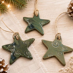 May include: Three star-shaped ceramic ornaments with a glossy dark green glaze and embossed spiral patterns. Each star features a hole at the top threaded with a natural jute twine loop for hanging, showcasing a smooth, reflective surface finish.