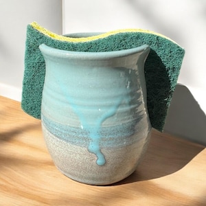 May include: A light blue ceramic sponge holder with a white and blue glaze. The holder is filled with a green and yellow sponge.