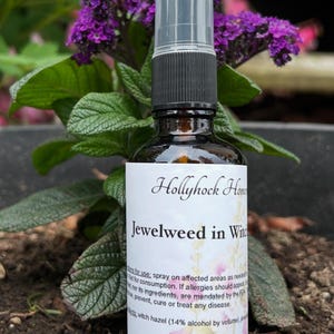 May include: A brown glass spray bottle with a black cap and clear spray nozzle. The label reads "Hollyhock Home Jewelweed in Witch Hazel". The bottle is surrounded by green leaves and purple flowers.