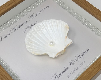 May include: A framed pearl wedding anniversary keepsake. The white seashell holds a single pearl. The text reads "Pearl Wedding Anniversary 30 Years" and "Brenda & Stephen 24 March 2020". The frame is light brown with a white background.
