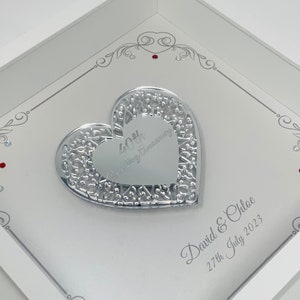 Può includere: Un regalo di anniversario incorniciato, bianco, a forma di cuore. Il cuore argentato presenta un design a filigrana aperta e le parole "40th Wedding Anniversary". I nomi "David & Chloe" e la data "27th July 2023" sono anch'essi visualizzati.