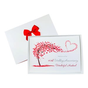 Puede incluir: Una caja de regalo rectangular blanca con un lazo rojo que contiene una tarjeta de felicitación. La tarjeta muestra un árbol con hojas rojas formando un corazón y el texto: With Love on Our 40th Wedding Anniversary Wonderful Husband en letra negra y roja.
